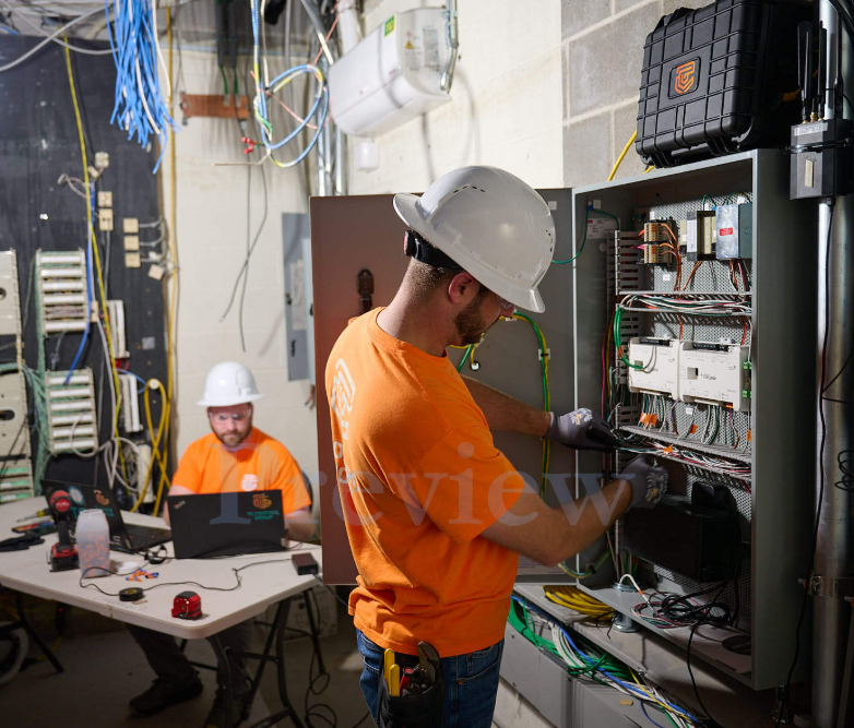 TC Controls technicians wiring a wall-mounted BMS controller while programming at a laptop in a mechanical room